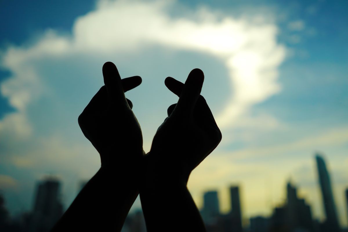 Silhouette hands making mini heart shaped hand gesture on blurred background of cityscape with high rise business metropolis building and skyscape with clouds at early morning or evening moment.